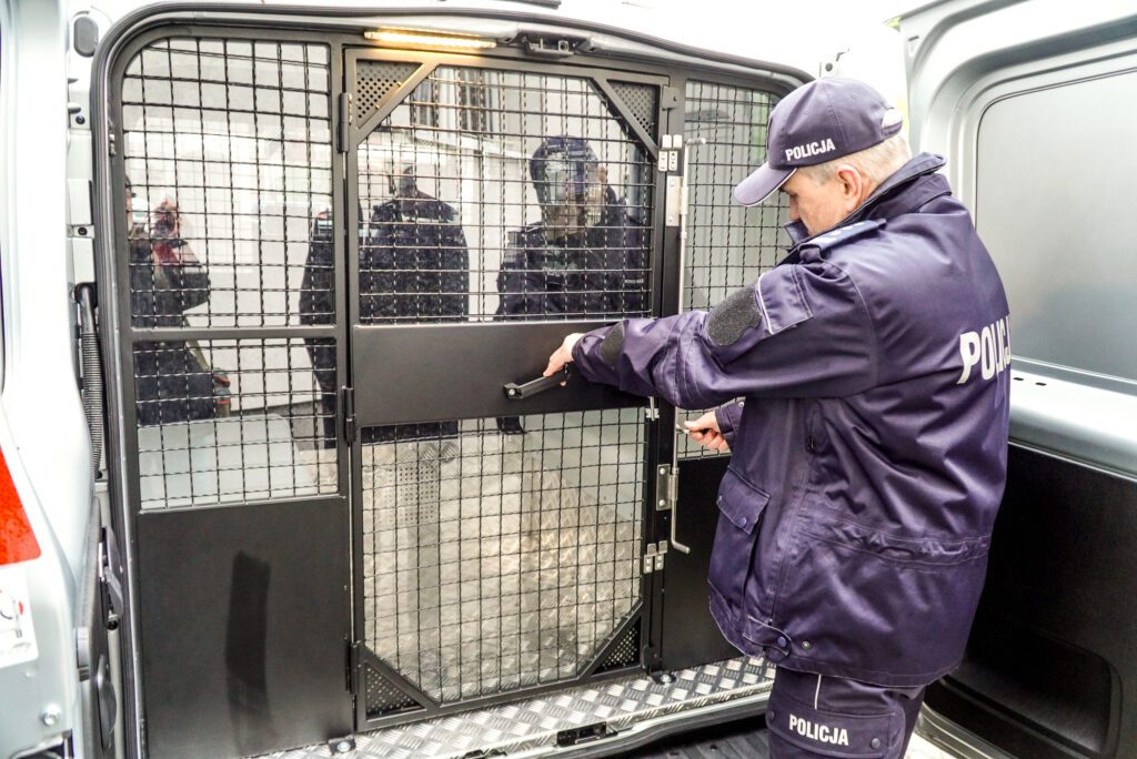 Policjant w mundurze i czapce z daszkiem otwiera czarną, metalową kratę wewnątrz tylnej części furgonetki. Przez otwarte drzwi widać wnętrze wyłożone blachą ryflowaną. Obok stoją fragmenty sylwetek innych osób w ciemnych płaszczach.