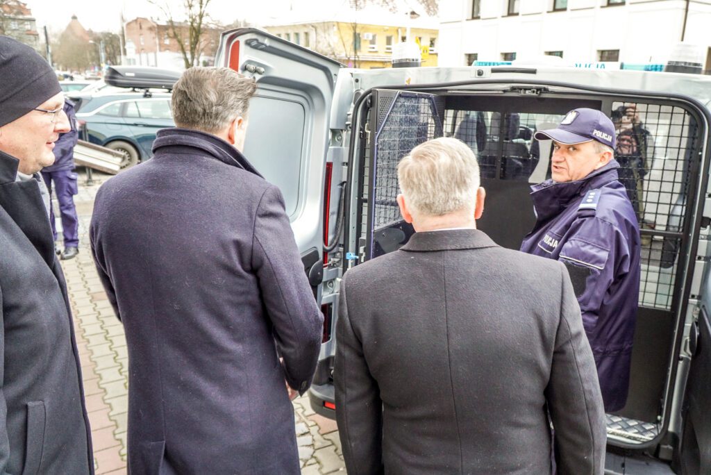 Grupa czterech mężczyzn przy otwartym tyle policyjnego vana. Trzech z nich, w cywilnych zimowych płaszczach, stoi tyłem lub bokiem do kamery. Czwarty, policjant w mundurze, stoi wewnątrz pojazdu i rozmawia z pozostałymi.