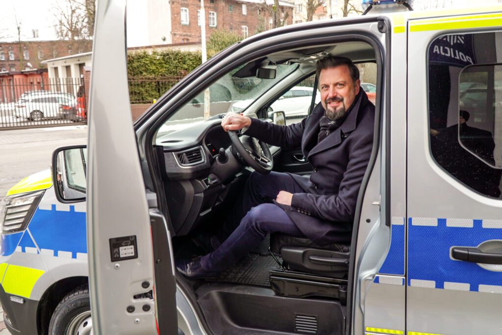 Mężczyzna siedzący na fotelu kierowcy w nowym oznakowanym radiowozie Policji marki Renault Trafic. Drzwi pojazdu są otwarte, a na zewnątrz widoczne są elementy zabudowy miejskiej. Samochód ma srebrne nadwozie z niebiesko-żółto-zielonymi pasami odblaskowymi oraz oznaczeniami „Policja”.