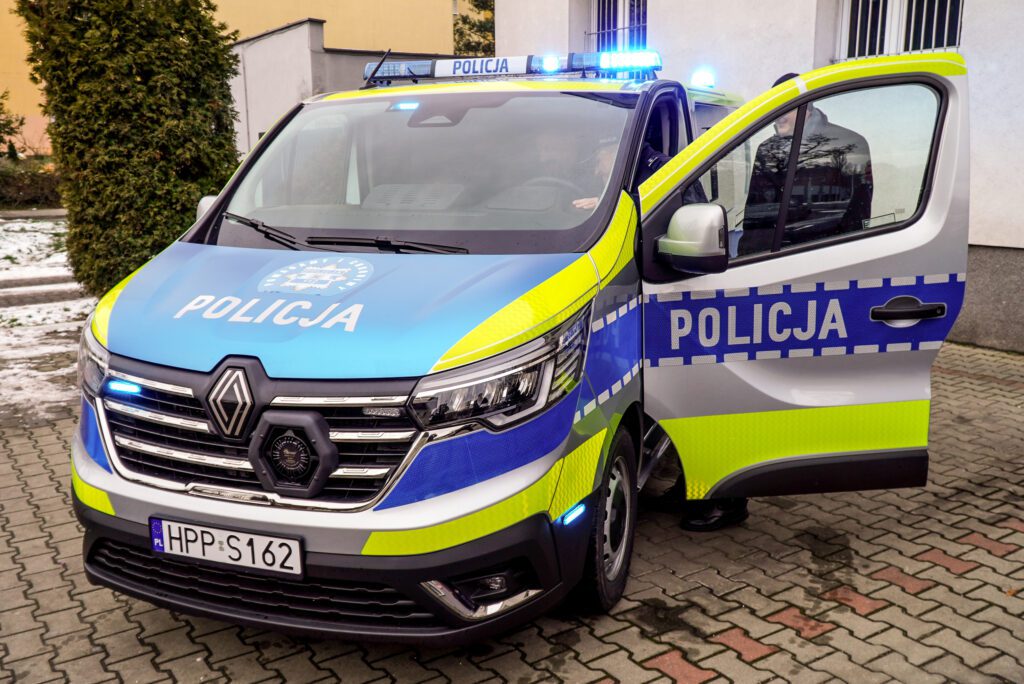 Widok z przodu na nowy radiowóz Policji zaparkowany przed budynkiem Komendy Miejskiej Policji. Na dachu pojazdu włączone są niebieskie sygnały świetlne. Na masce i drzwiach widoczne oznaczenia „Policja”, a na nadwoziu fluorescencyjne pasy poprawiające widoczność.