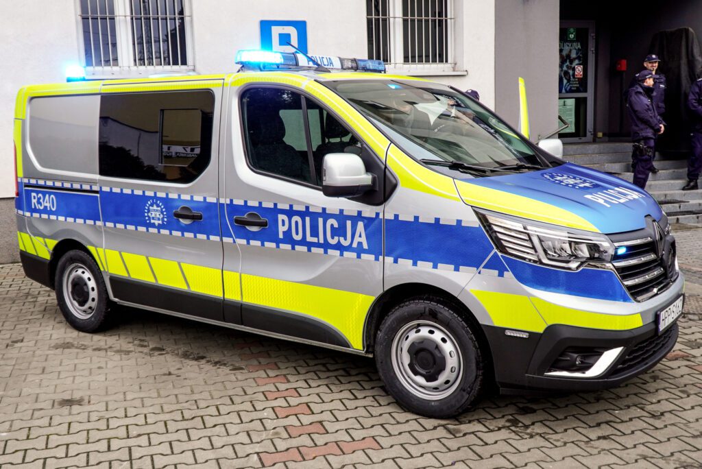 Boczny widok oznakowanego radiowozu Renault Trafic zaparkowanego na dziedzińcu komendy. Samochód ma charakterystyczne niebieskie oznaczenia policyjne, jaskrawe pasy odblaskowe oraz włączone światła ostrzegawcze. W tle widoczny budynek Policji oraz funkcjonariusze.