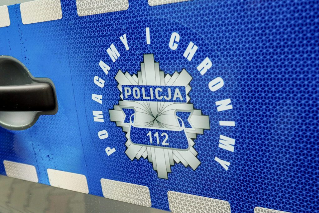 Zbliżenie na herb Policji umieszczony na bocznych drzwiach radiowozu. Na niebieskim tle wokół herbu widoczny napis „Pomagamy i chronimy”, a w środku herbu napis „Policja” oraz numer alarmowy 112. Faktura odblaskowej folii podkreśla funkcję bezpieczeństwa pojazdu.