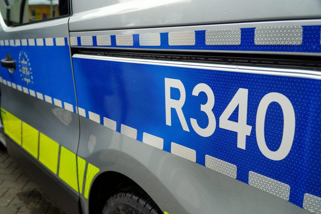 Detal bocznej części radiowozu z widocznym oznaczeniem „R340” umieszczonym na niebieskim pasie odblaskowym. W tle fragment karoserii oraz koło pojazdu zaparkowanego na utwardzonym placu.