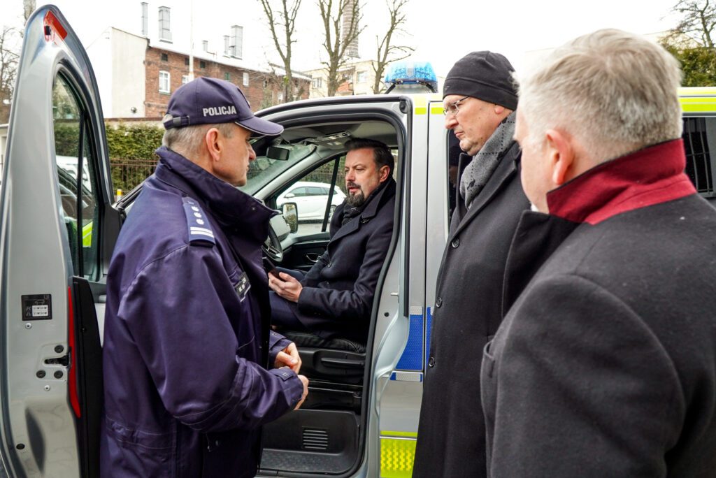 Kilku mężczyzn stoi przy otwartych drzwiach radiowozu Policji. Jeden z nich siedzi na miejscu kierowcy, pozostali – w tym funkcjonariusz w mundurze – rozmawiają obok pojazdu. Scena odbywa się przed budynkiem komendy.