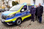 Grupa osób stojących obok nowego radiowozu Policji zaparkowanego przed komendą. Wśród nich funkcjonariusz w mundurze oraz przedstawiciele władz miasta. Na dachu pojazdu włączone są niebieskie sygnały świetlne, a wszyscy pozują do wspólnego zdjęcia.
