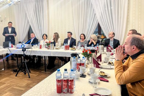 Uczestnicy posiedzenia Rady Gospodarczej siedzą przy długim stole w eleganckiej sali Zameczku w Michałkowicach. Po lewej stronie stoi przewodniczący Rady, który czyta dokumenty, rozpoczynając obrady. Na stole widoczne są filiżanki, woda, napoje oraz drobny poczęstunek. Uczestnicy skupieni są na wystąpieniu, a w tle widać jasne zasłony i klasyczne oświetlenie sali.