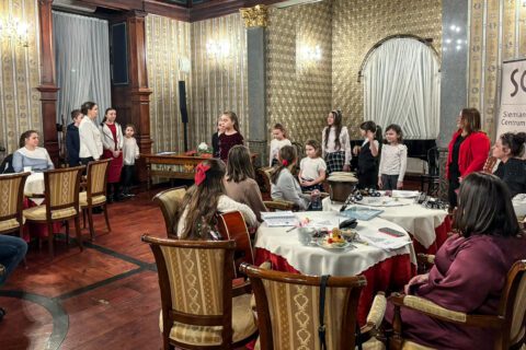 Widok na część sali, gdzie przy stolikach siedzą zaproszeni goście – kobiety i mężczyźni w różnym wieku. Przed nimi leżą śpiewniki i filiżanki. Uczestnicy słuchają występu i rozmawiają w przerwach między kolędami.