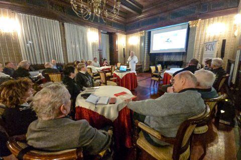 Szeroki plan sali w SCK Willa Fitznera z seniorami zajmującymi miejsca przy stolikach i prowadzącą prezentującą informacje o naborze do Rady Seniorów.