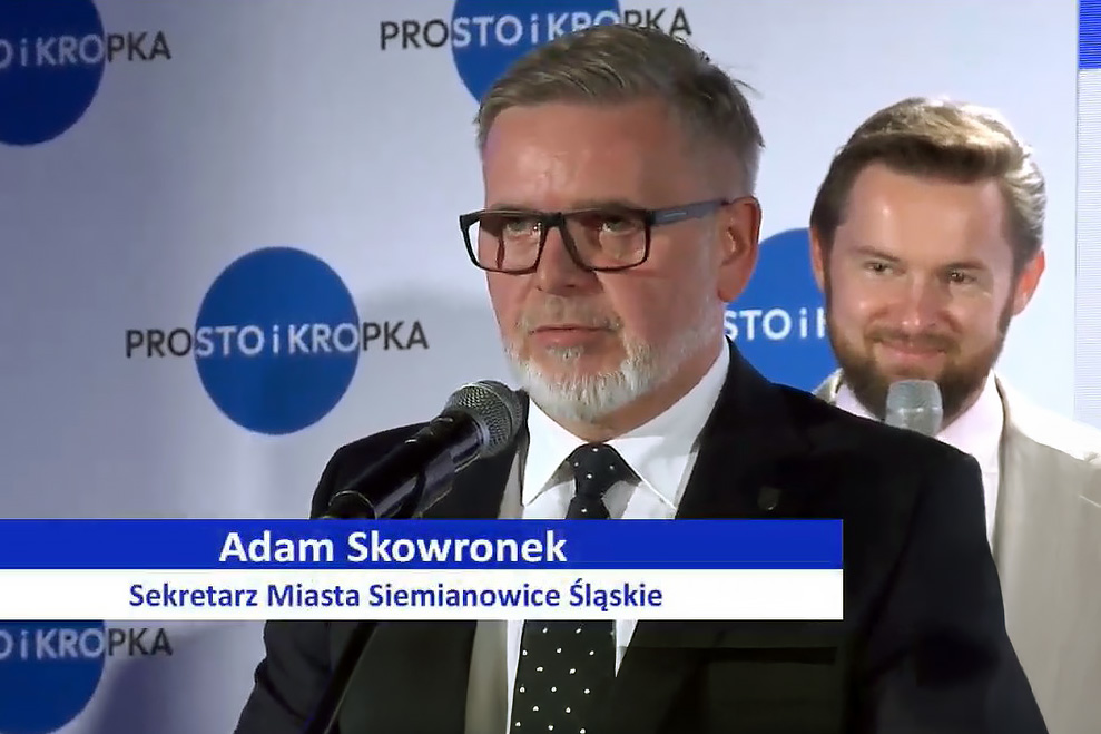 Adam Skowronek, Sekretarz Miasta Siemianowice Śląskie, przemawia do mikrofonu podczas oficjalnego wydarzenia. W tle widoczne logo inicjatywy „Prosto i kropka”. Zdjęcie wykonane w trakcie uroczystości podpisania Deklaracji Prostego Języka.