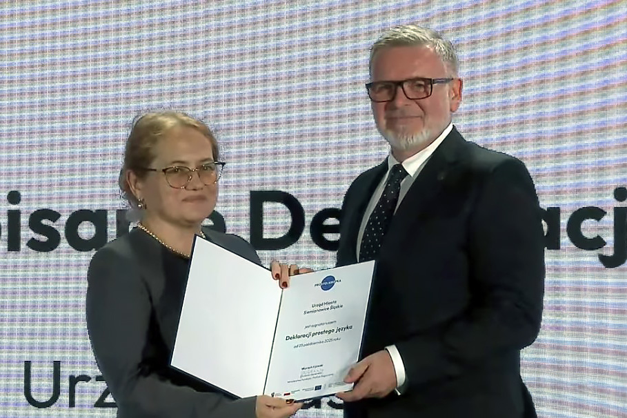 Sekretarz Miasta Siemianowice Śląskie Adam Skowronek stoi obok przedstawicielki organizatorów i prezentuje otwarty dokument Deklaracji Prostego Języka. Oboje pozują do zdjęcia na tle ekranu wydarzenia.