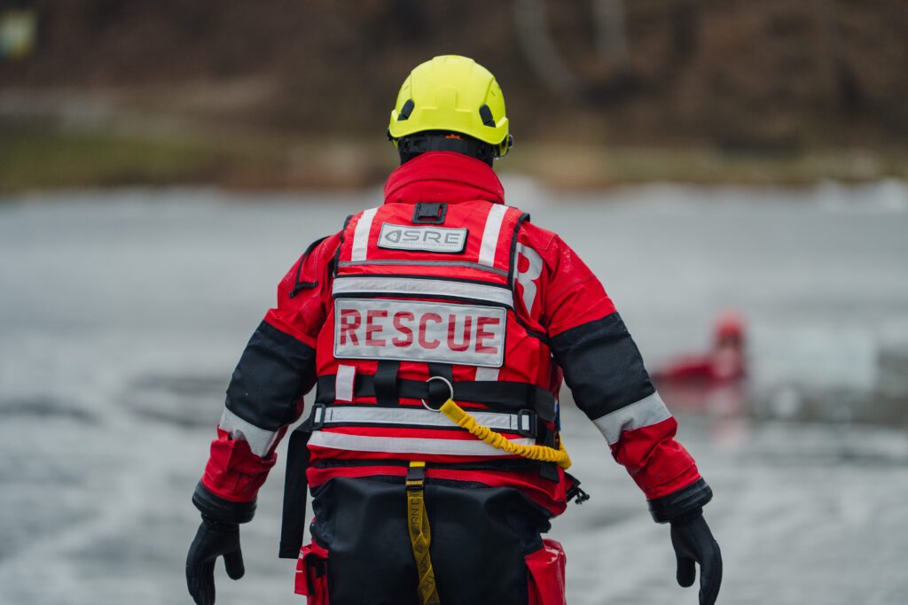 Ratownik w czerwonym kombinezonie z napisem „RESCUE” na plecach, w kasku i kamizelce asekuracyjnej, stoi w wodzie podczas ćwiczeń ratownictwa lodowego. W tle widać osobę w wodzie oraz zamarznięty akwen.