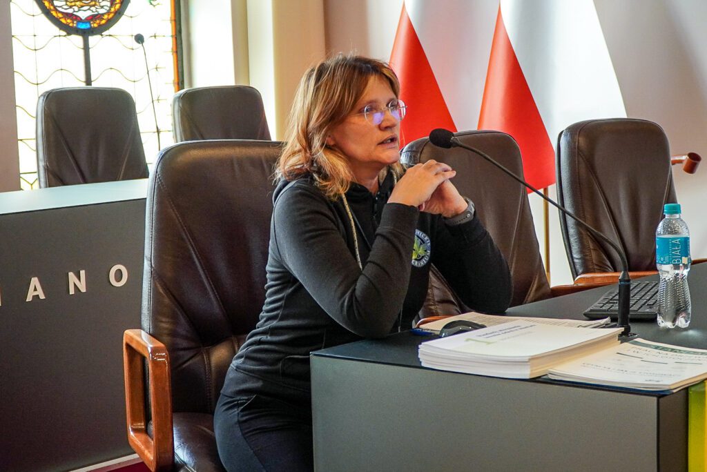 Kobieta w okularach siedzi przy stole prezydialnym w sali posiedzeń ratusza i przemawia do zgromadzonych przez mikrofon. Przed nią leżą dokumenty, a w tle widoczne są flagi oraz elementy wystroju sali obrad.