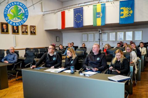 Szerokie ujęcie sali posiedzeń, w której przy długich stołach siedzą uczestnicy spotkania. Osoby słuchają prelegentów, mają przed sobą dokumenty i notatki. Na ścianach widoczne są flagi i portrety.