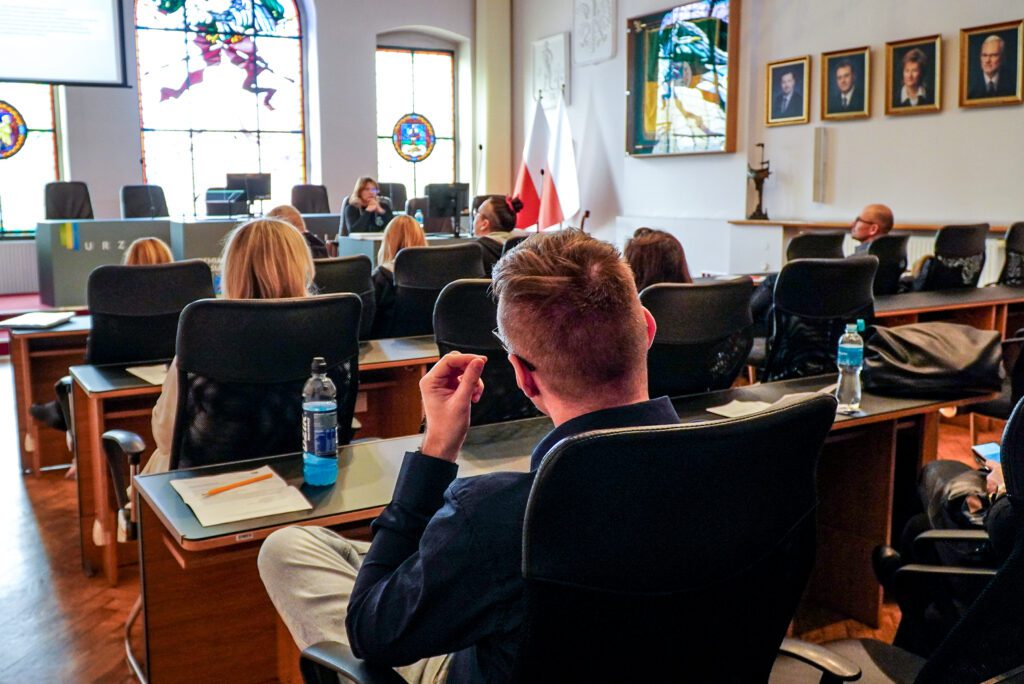 Uczestnicy spotkania siedzą w rzędach w sali obrad, skupieni na omawianych zagadnieniach. Na stołach znajdują się teczki, dokumenty oraz urządzenia mobilne wykorzystywane do notowania.