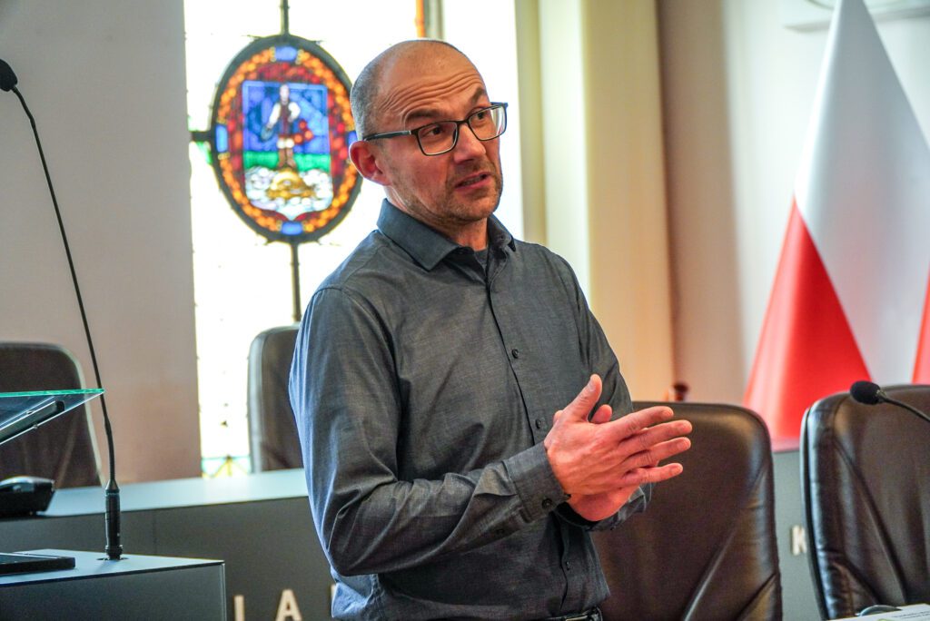 Mężczyzna stojący w sali posiedzeń ratusza przemawia do zgromadzonych. Gestykuluje dłońmi, wyjaśniając omawiane zagadnienia. W tle widoczne są witraże oraz elementy wyposażenia sali obrad.
