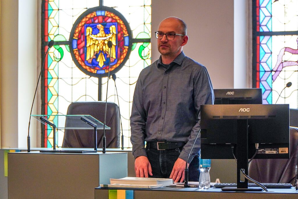 Mężczyzna stoi przy mównicy w sali obrad, opierając dłonie na dokumentach. Za nim widoczny jest witraż oraz monitory komputerowe. Ujęcie podkreśla formalny charakter wystąpienia.