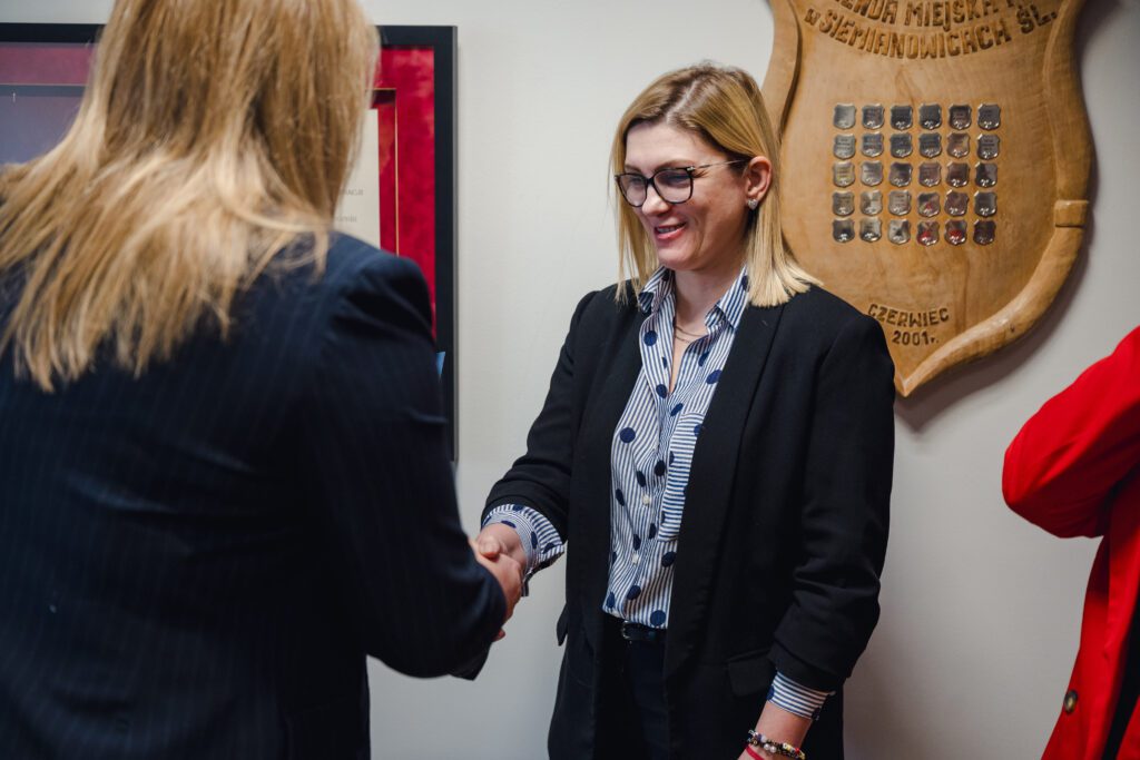 Kobieta w ciemnym żakiecie (widoczna od tyłu) podaje rękę uśmiechniętej kobiecie w ciemnej marynarce i okularach. W tle ozdobna tarcza z metalowymi plakietkami.