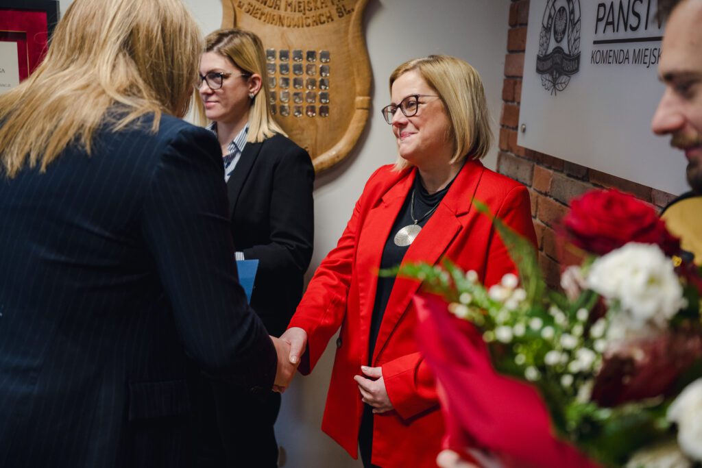 Beata Gałecka (widoczna z lewej strony tyłem) podaje rękę kobiecie w czerwonej marynarce. W tle widać drugą pracownicę oraz tablicę pamiątkową na ścianie.
