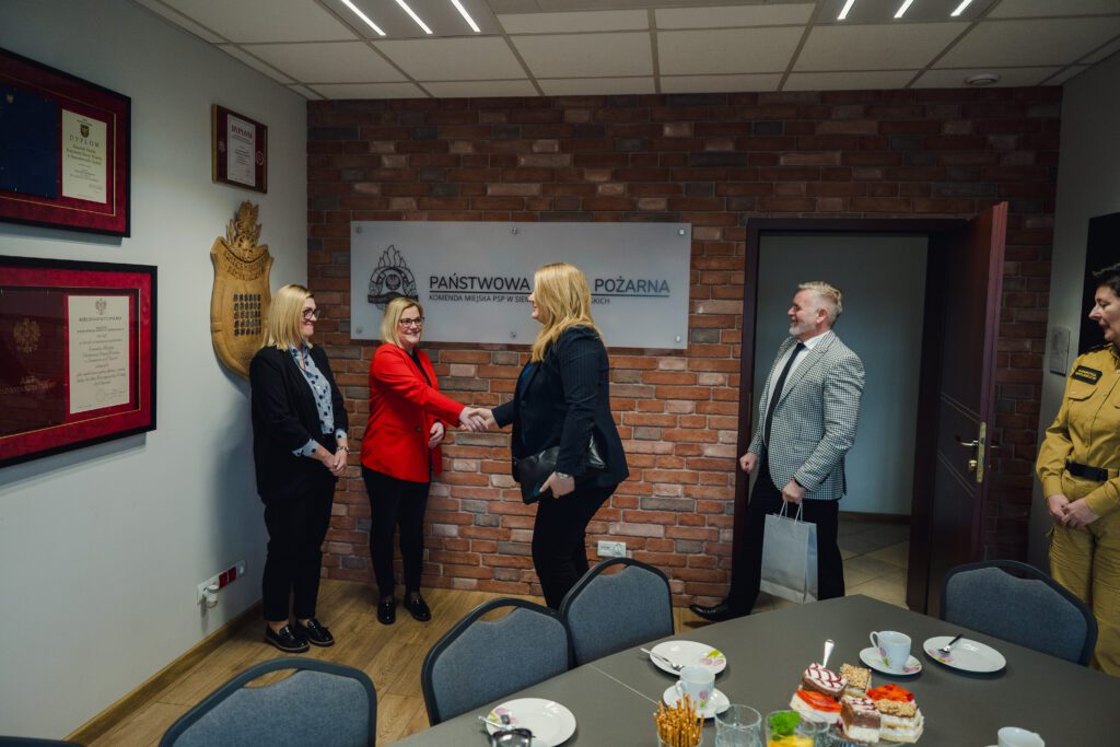 Trzy kobiety stoją przy ścianie z tablicą „Państwowa Straż Pożarna – Komenda Miejska PSP w Siemianowicach Śląskich”. Dwie z nich podają sobie ręce na powitanie. Obok stoi mężczyzna z torbą prezentową oraz funkcjonariuszka w mundurze. Na pierwszym planie widać stół z filiżankami i ciastem.