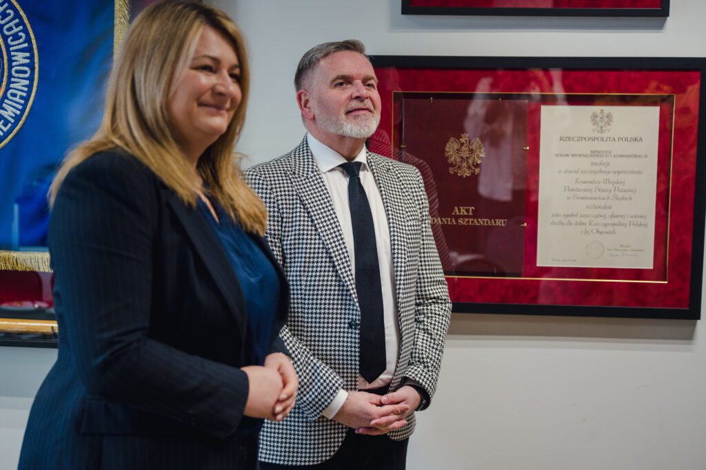 Kobieta w ciemnej marynarce i mężczyzna w marynarce w drobną kratkę stoją obok siebie w pomieszczeniu biurowym. W tle na ścianie widać oprawiony w ramę akt nadania sztandaru.
