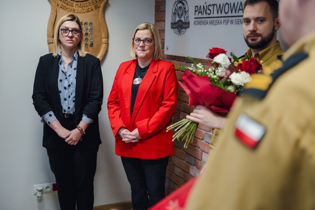 Dwie kobiety stojące pod ścianą w biurze. Jedna w ciemnym żakiecie i koszuli w kropki, druga w czerwonej marynarce. Z prawej strony widać fragment munduru strażackiego i bukiet czerwonych róż.