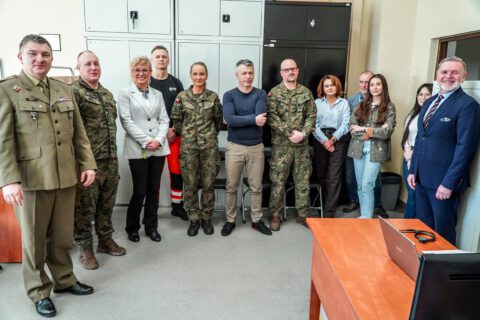 Grupa osób stoi w biurze. Oficer w mundurze polowym (ppłk Krzysztof Szmyt) przemawia do urzędników i żołnierzy. Po prawej Sekretarz Miasta Adam Skowronek. W tle białe szafy