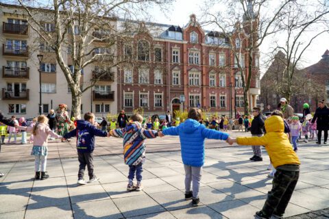 Dzieci trzymają się za ręce i tworzą duże koło na placu. W tle ceglany, historyczny budynek.
