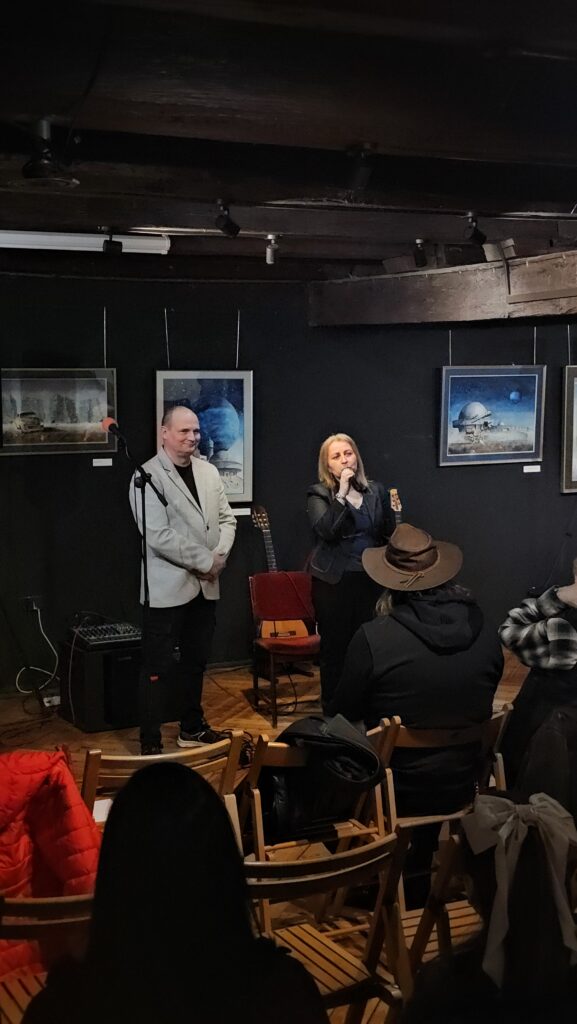 (1 dzień festiwalu – Otwarcie) Mężczyzna w jasnej marynarce oraz kobieta trzymająca mikrofon stoją przed siedzącą publicznością. Tło stanowią ciemne ściany z zawieszonymi obrazami. Wnętrze ma charakter kameralnej galerii.