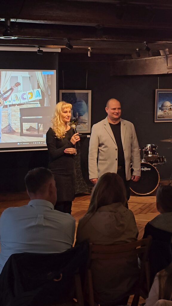 (2 dzień festiwalu – Koncert wychowanków MDK im. dr. H. Jordana) Kobieta z blond włosami trzyma mikrofon i przemawia obok mężczyzny w jasnej marynarce. Stoją na drewnianym parkiecie przed ekranem projekcyjnym. W tle widać instrumenty muzyczne oraz obrazy na ciemnej ścianie.