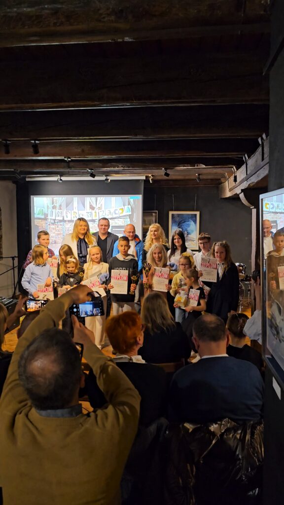 (2 dzień festiwalu – Koncert wychowanków MDK im. dr. H. Jordana) Zdjęcie grupowe wszystkich uczestników i organizatorów na scenie. Dzieci trzymają pamiątkowe dyplomy i statuetki. W dolnej części kadru widać osoby z publiczności fotografujące grupę telefonami