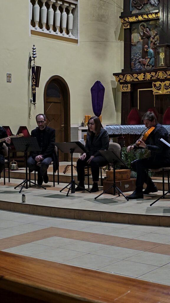 (3 dzień festiwalu – Koncert Muzyki dawnej – zespół SCANDICUS, kościół pw. Św. Antoniego) Zespół muzyczny wykonuje utwór na podwyższeniu przed ołtarzem. Widoczna jest kobieta śpiewająca z nut oraz mężczyzna grający na skrzypcach. Wnętrze charakteryzuje się jasnymi ścianami i klasyczną architekturą.