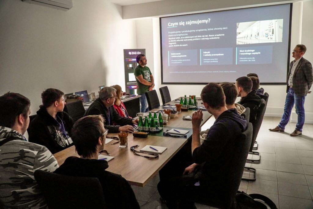 Uczniowie siedzą przy stole konferencyjnym i słuchają prezentacji o działalności firmy wyświetlanej na ekranie.