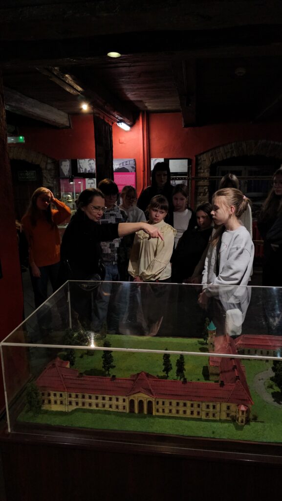 (1 dzień festiwalu – zwiedzanie grupy plastycznej „Okienko”) Grupa młodych ludzi stoi wokół szklanej gabloty z dużą makietą przedstawiającą klasycystyczny budynek o czerwonym dachu. Kobieta w okularach wskazuje palcem na konkretny element makiety podczas objaśniania wystawy.