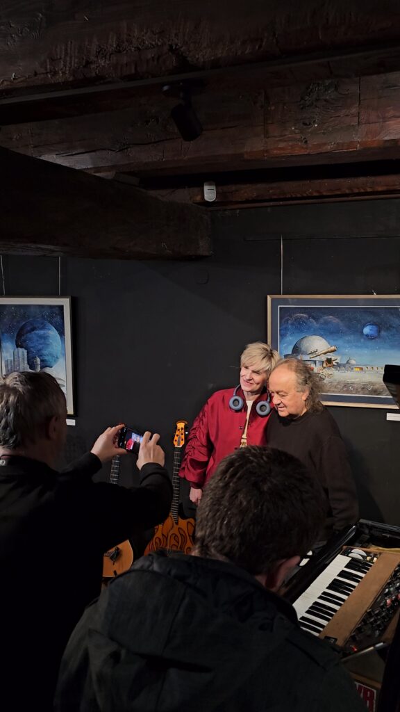 (1 dzień festiwalu – Koncert Józef Skrzek i Adam Bul) Kobieta w czerwonej kurtce z niebieskimi słuchawkami na szyi pozuje do zdjęcia z mężczyzną. Przed nimi stoi osoba, która rejestruje tę chwilę za pomocą smartfona. W tle widać fragment fortepianu oraz obrazy na ścianie.