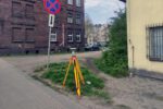 Sprzęt geodezyjny ustawiony na statywie przy chodniku obok budynku mieszkalnego i znaku zakazu zatrzymywania się. Zdjęcie ilustruje ogłoszenie dotyczące kradzieży urządzenia pomiarowego wykorzystywanego przez geodetów.