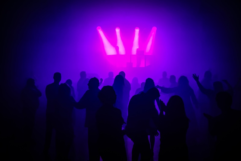 Zdjęcie przedstawia grupę ludzi tańczących na parkiecie podczas Dyskoteki 40-latków. Postacie widoczne są jako ciemne sylwetki na tle intensywnego, kolorowego oświetlenia scenicznego w odcieniach fioletu, różu i niebieskiego. W górnej części obrazu znajdują się jasne reflektory skierowane w stronę parkietu, częściowo rozproszone przez dym sceniczny, co tworzy miękką, rozświetloną poświatę. Na pierwszym planie widoczne są sylwetki osób w różnych pozach tanecznych – niektóre trzymają ręce uniesione do góry, inne stoją blisko siebie, sugerując rozmowę lub wspólny taniec.