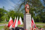Pomnik św. Floriana ustawiony na kamiennym postumencie, wokół którego znajdują się biało-czerwone flagi oraz złożone wieńce i kwiaty. Na cokole widoczny jest napis upamiętniający patrona i rocznicę miasta, a całość otoczona jest zielenią parku.