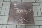 Metalowa, kwadratowa pamiątkowa płyta wmurowana w chodnik z kostki brukowej. Na płycie znajduje się duża gwiazda z logo zespołu SBB w środku, herb Katowic, logo "Kato Blues Faces" oraz kod QR.