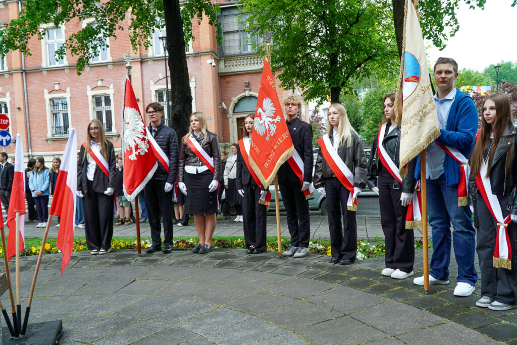 Młodzież szkolna stojąca w pocztach sztandarowych podczas uroczystości Narodowego Dnia Zwycięstwa. Uczniowie mają biało-czerwone szarfy i trzymają sztandary przed budynkiem w centrum miasta.