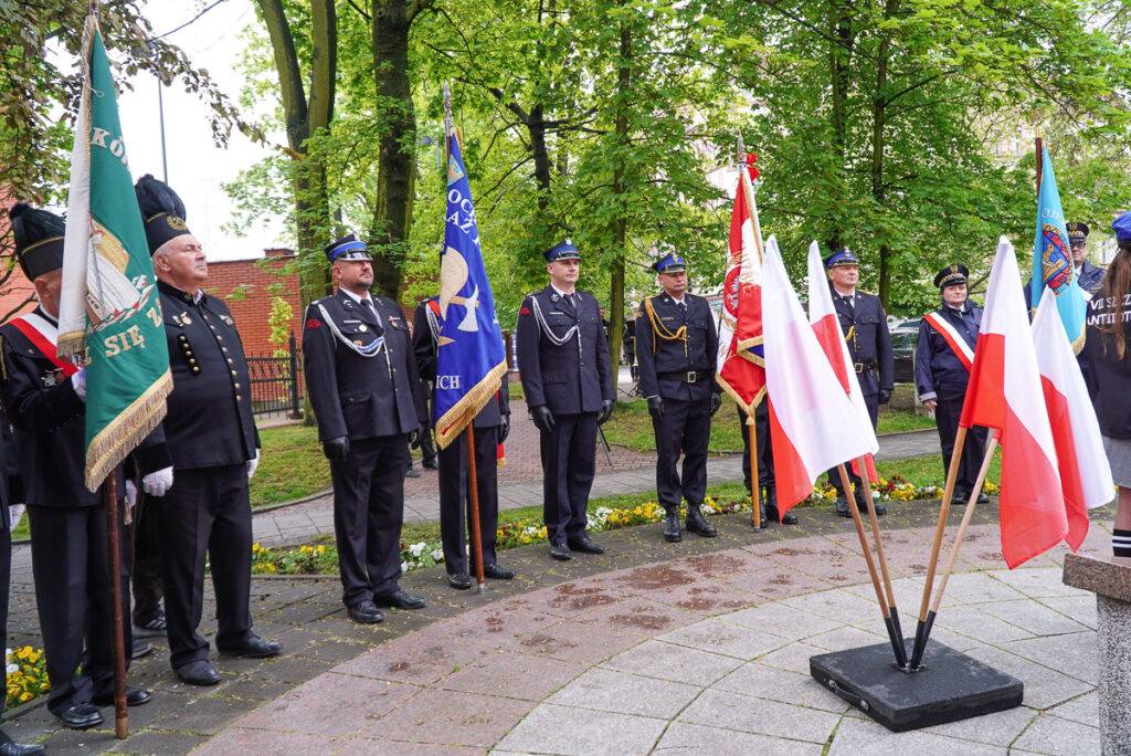 Strażacy w galowych mundurach oraz przedstawiciele pocztów sztandarowych stoją podczas obchodów Narodowego Dnia Zwycięstwa. Na pierwszym planie widoczne są biało-czerwone flagi ustawione przy pomniku.