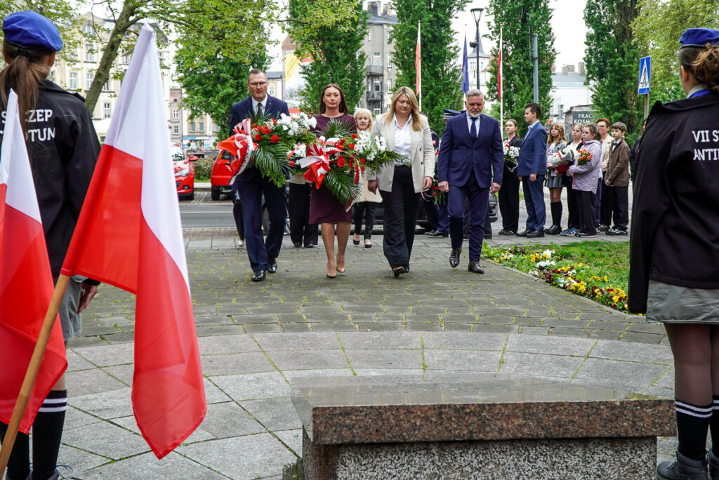 Przedstawiciele władz i delegacji przechodzą alejką w kierunku pomnika, niosąc biało-czerwone wiązanki kwiatów. Po obu stronach stoją harcerze oraz uczestnicy uroczystości.