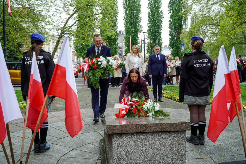 Kobieta składa biało-czerwoną wiązankę kwiatów pod pomnikiem podczas obchodów Narodowego Dnia Zwycięstwa. W tle stoją przedstawiciele władz miasta i uczestnicy uroczystości.