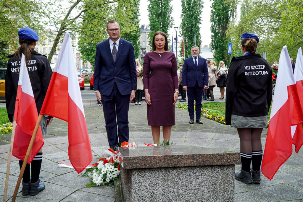 Przedstawiciele władz miasta stoją w ciszy przed pomnikiem po złożeniu kwiatów. Po bokach widoczne są biało-czerwone flagi oraz harcerze pełniący wartę honorową.