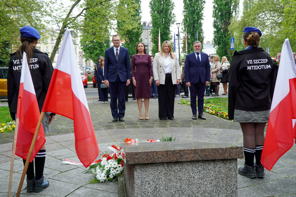 Przedstawiciele władz miasta oraz uczestnicy uroczystości stoją przed pomnikiem podczas obchodów Narodowego Dnia Zwycięstwa. U stóp pomnika leżą biało-czerwone wiązanki kwiatów, a wokół zgromadzili się mieszkańcy i młodzież.