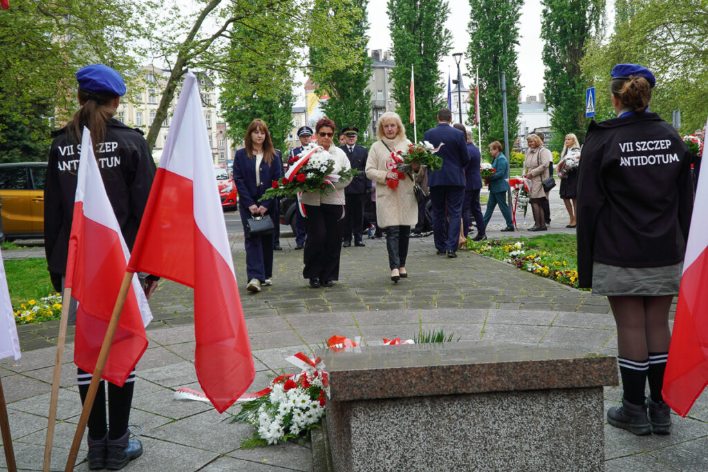 Uczestnicy uroczystości niosą biało-czerwone wiązanki kwiatów w kierunku pomnika. Po obu stronach alejki stoją harcerze oraz ustawione są polskie flagi.