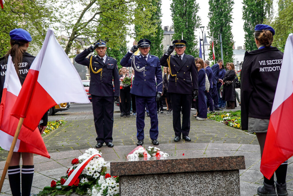 Trzech przedstawicieli służb mundurowych stoi na baczność i salutuje przed pomnikiem podczas obchodów Narodowego Dnia Zwycięstwa. U stóp pomnika leżą biało-czerwone kwiaty i wieńce.