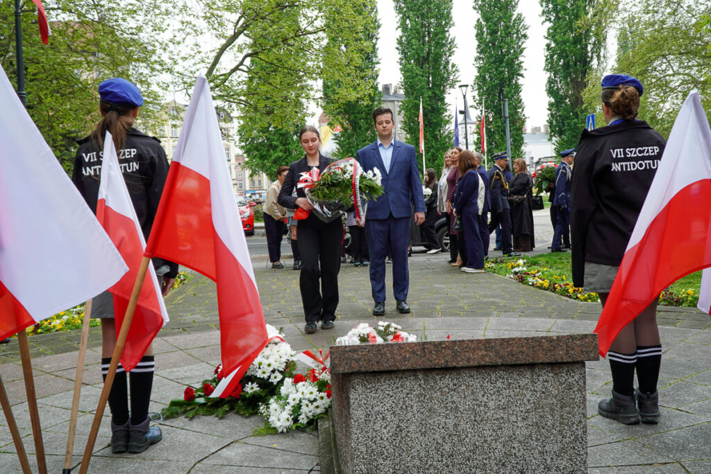 Dwójka młodych uczestników uroczystości niesie wiązankę kwiatów w stronę pomnika podczas obchodów Narodowego Dnia Zwycięstwa. W tle widoczni są pozostali uczestnicy wydarzenia.