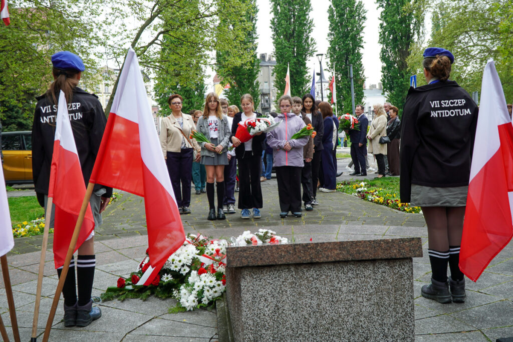 Grupa dzieci, młodzieży i dorosłych uczestników uroczystości stoi przed pomnikiem z kwiatami w rękach. Po bokach alejki widoczne są biało-czerwone flagi oraz harcerze pełniący wartę.