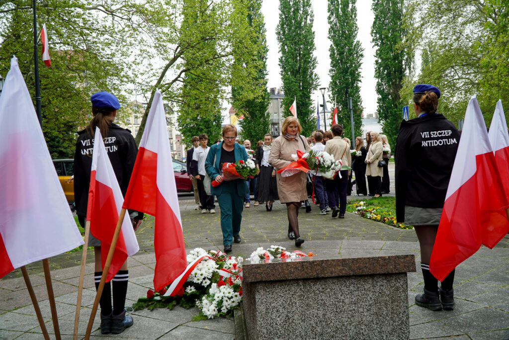 Uczestnicy obchodów Narodowego Dnia Zwycięstwa podchodzą z wiązankami kwiatów do pomnika. W tle zgromadzili się mieszkańcy oraz przedstawiciele różnych środowisk i organizacji.