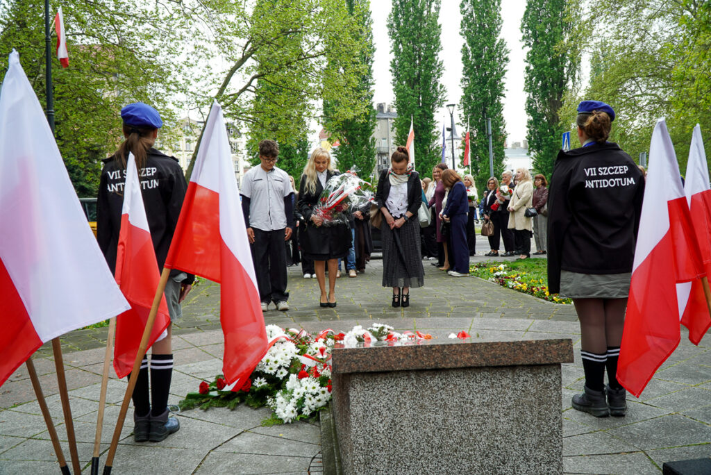 Uczestnicy uroczystości stoją w ciszy przed pomnikiem po złożeniu biało-czerwonych wiązanek kwiatów. Po bokach stoją harcerze oraz ustawione są polskie flagi.